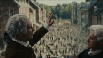 "Two men in 18th-century attire stand on a balcony overlooking a large crowd filling a colonial street lined with brick buildings, with trees visible in the background, suggesting a historical event or public gathering."